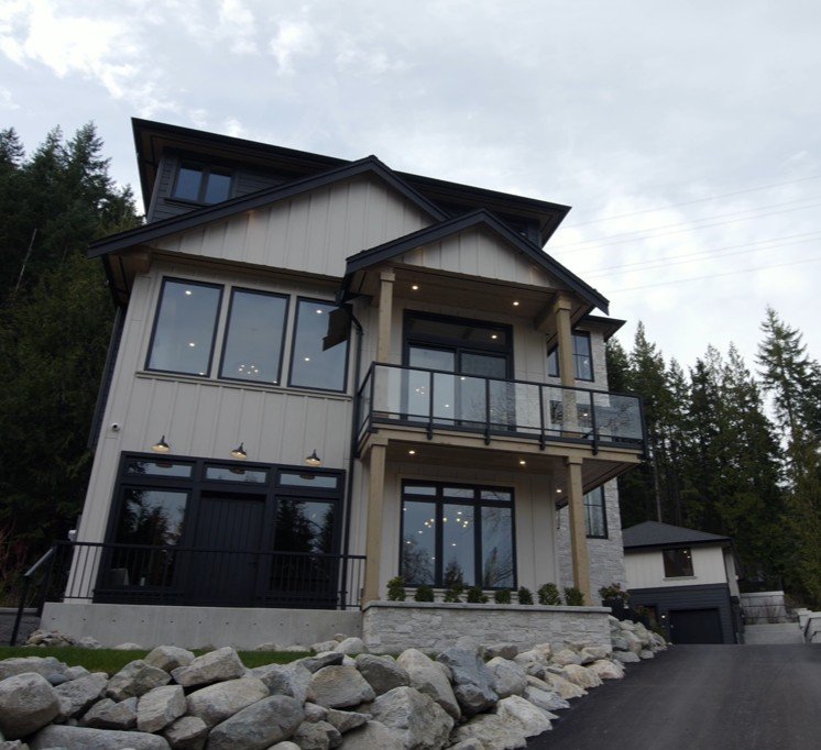 Modern multi-story house with large windows, a balcony with glass railing, and landscaped rocks in front, situated among trees.