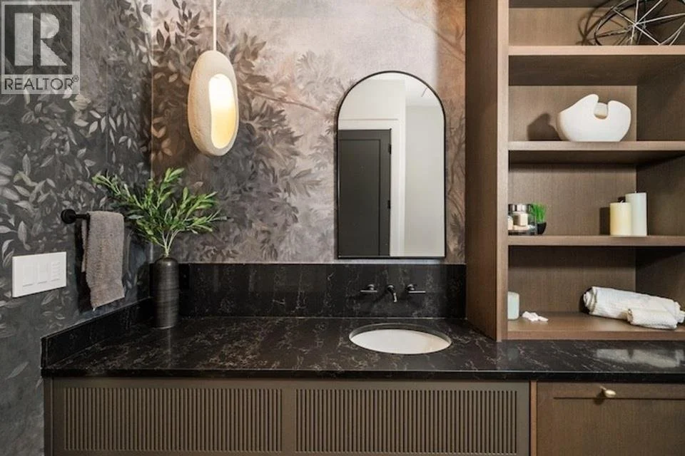 Modern bathroom vanity with black marble countertop, oval mirror, decorative vase with green plant, wall-mounted tap, and open wooden shelves with decorative items and towels.