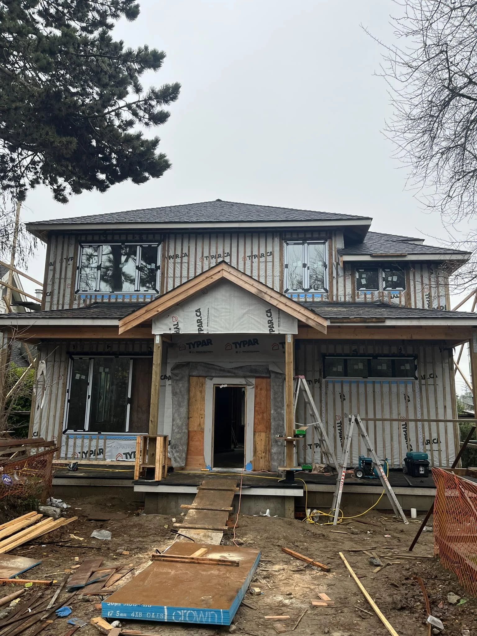Two-story house under construction with plywood framing, pitched roof, and large windows, surrounded by construction tools and materials.