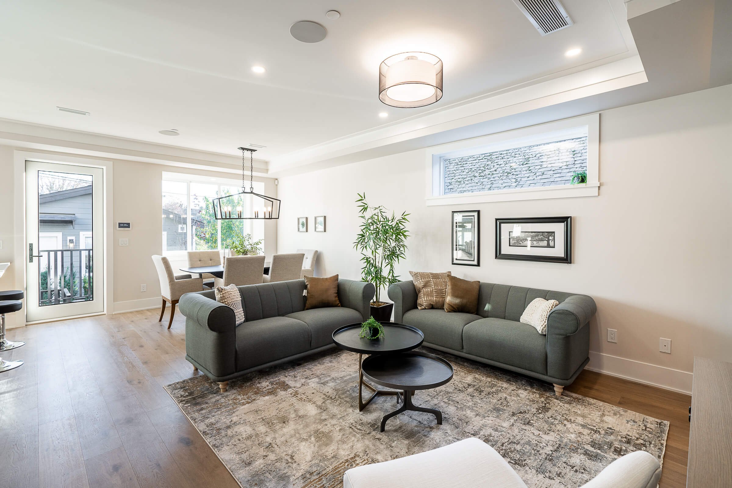 Bright open living room with two gray sofas, decorative pillows, and a round black coffee table on an area rug. Behind the sofas is a dining area with a table and six chairs, and a window with a view of neighboring houses. The walls are decorated wit