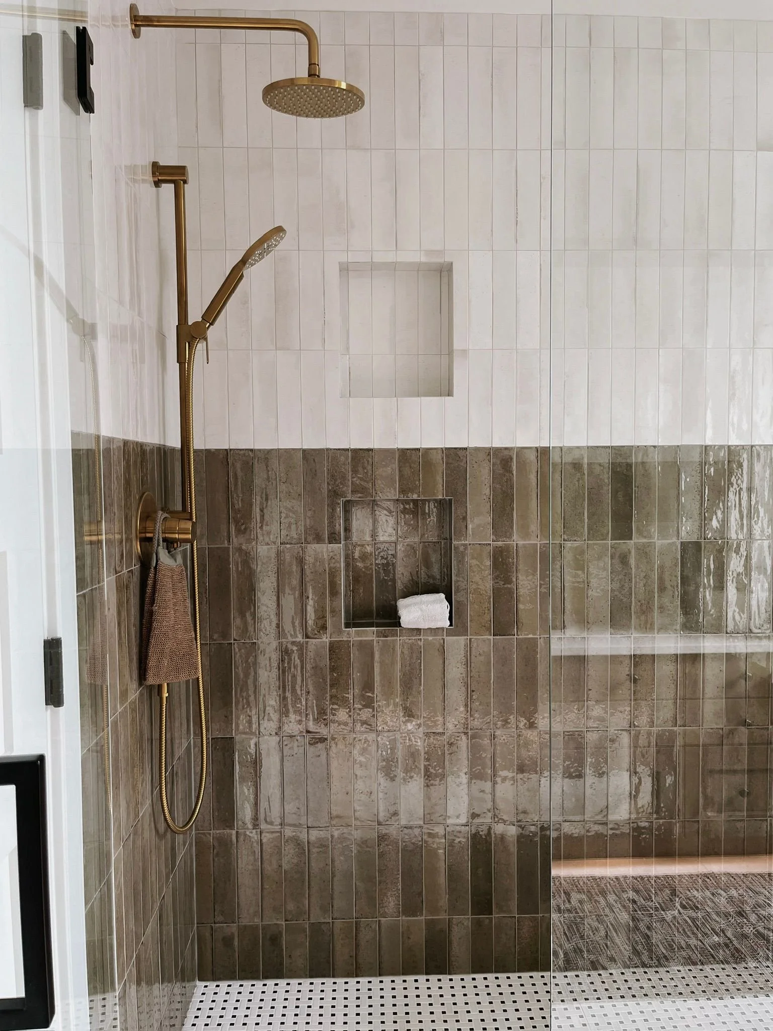 A modern shower area with a dual showerhead system and brown tiled walls. The shower has one large overhead rain shower and a separate handheld shower. There are two built-in niches, one with a rolled white towel. The floor has small white tiles with black dots, and the shower area is enclosed with a clear glass door.