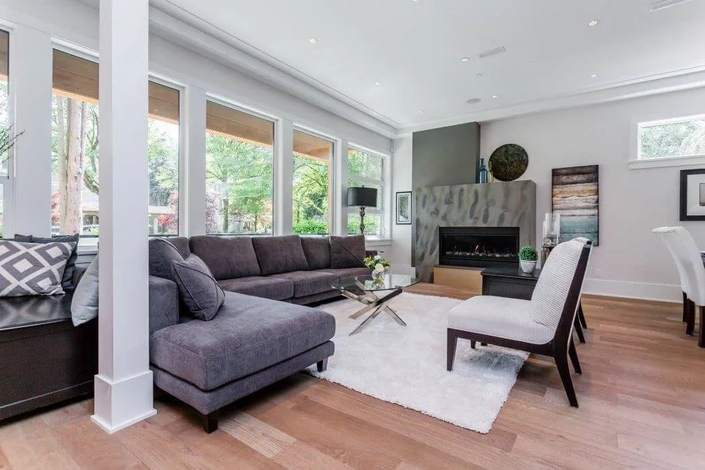 Living room with large gray sectional sofa, white armchairs, glass coffee table, fireplace, and wide windows overlooking a green outside view.