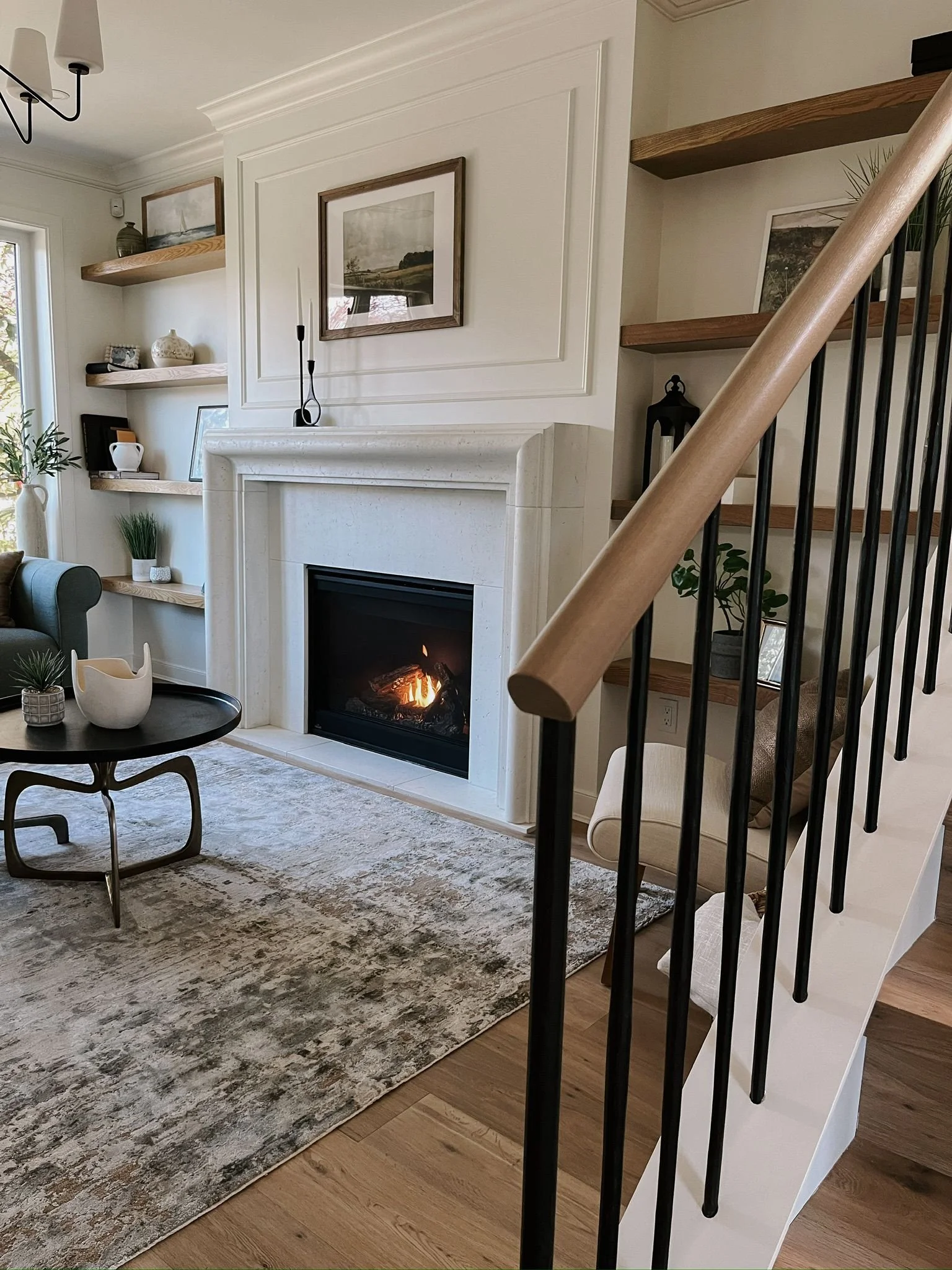 Living room with a white fireplace, a black coffee table, a gray rug, built-in wooden shelves with decorative items, a framed landscape picture above the fireplace, and a staircase with a wooden handrail and black balusters.