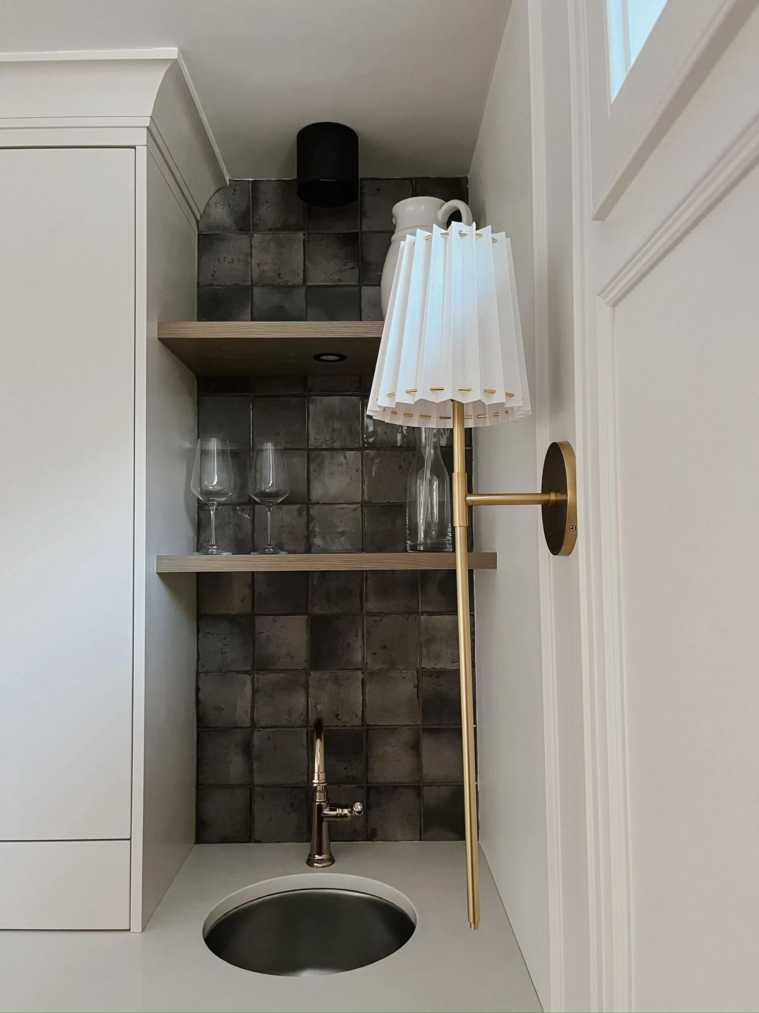 Interior of a small bar area with dark tiled backsplash, two shelves holding wine glasses, a pitcher and a black container, a small sink with a brass faucet, a white wall-mounted lamp with a pleated shade, and a white cabinet.