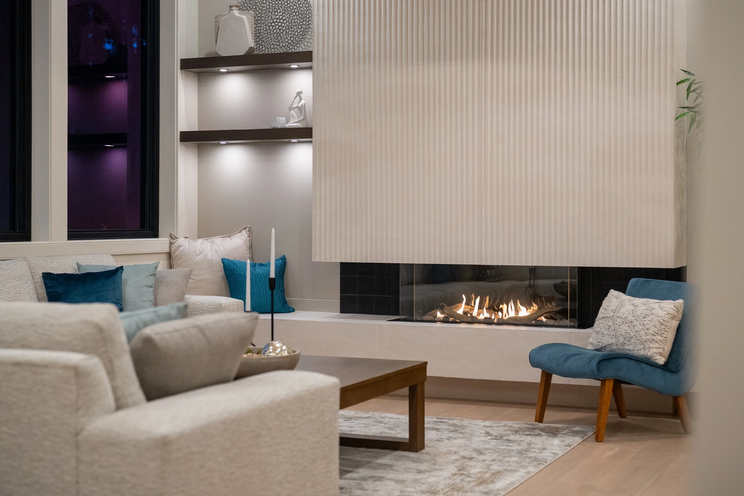 Modern living room with a fireplace, beige sofa with blue and white pillows, blue armchair with a patterned cushion, a wooden coffee table with a candle, and built-in bookshelves with decorative items, illuminated by soft lighting.