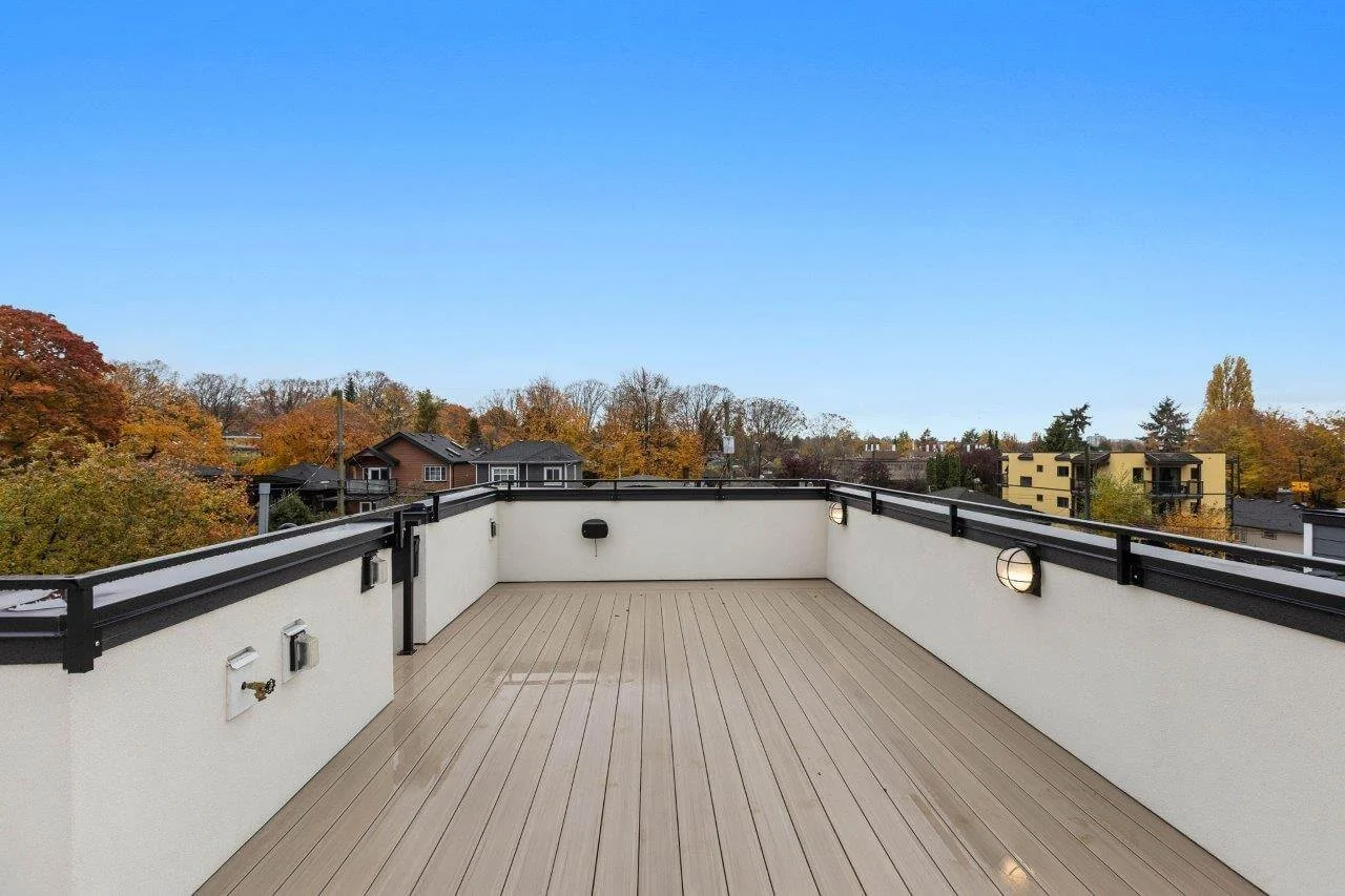 Empty rooftop terrace with beige wooden flooring, white walls, black railing, and view of neighborhood with trees and houses under a clear blue sky.