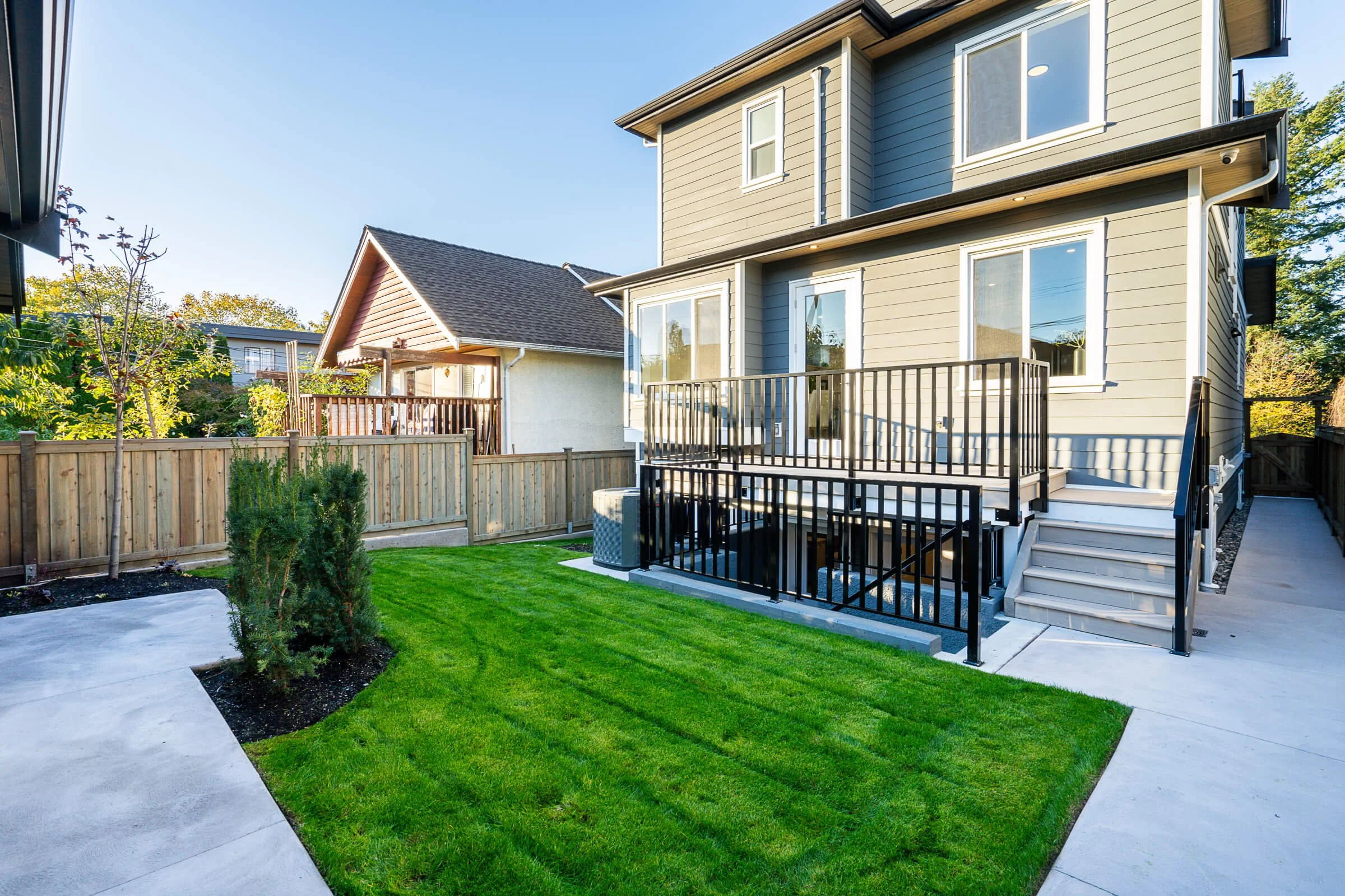 Backyard with green lawn, small trees, wooden fence, concrete pathway, and house with raised deck and black railing.