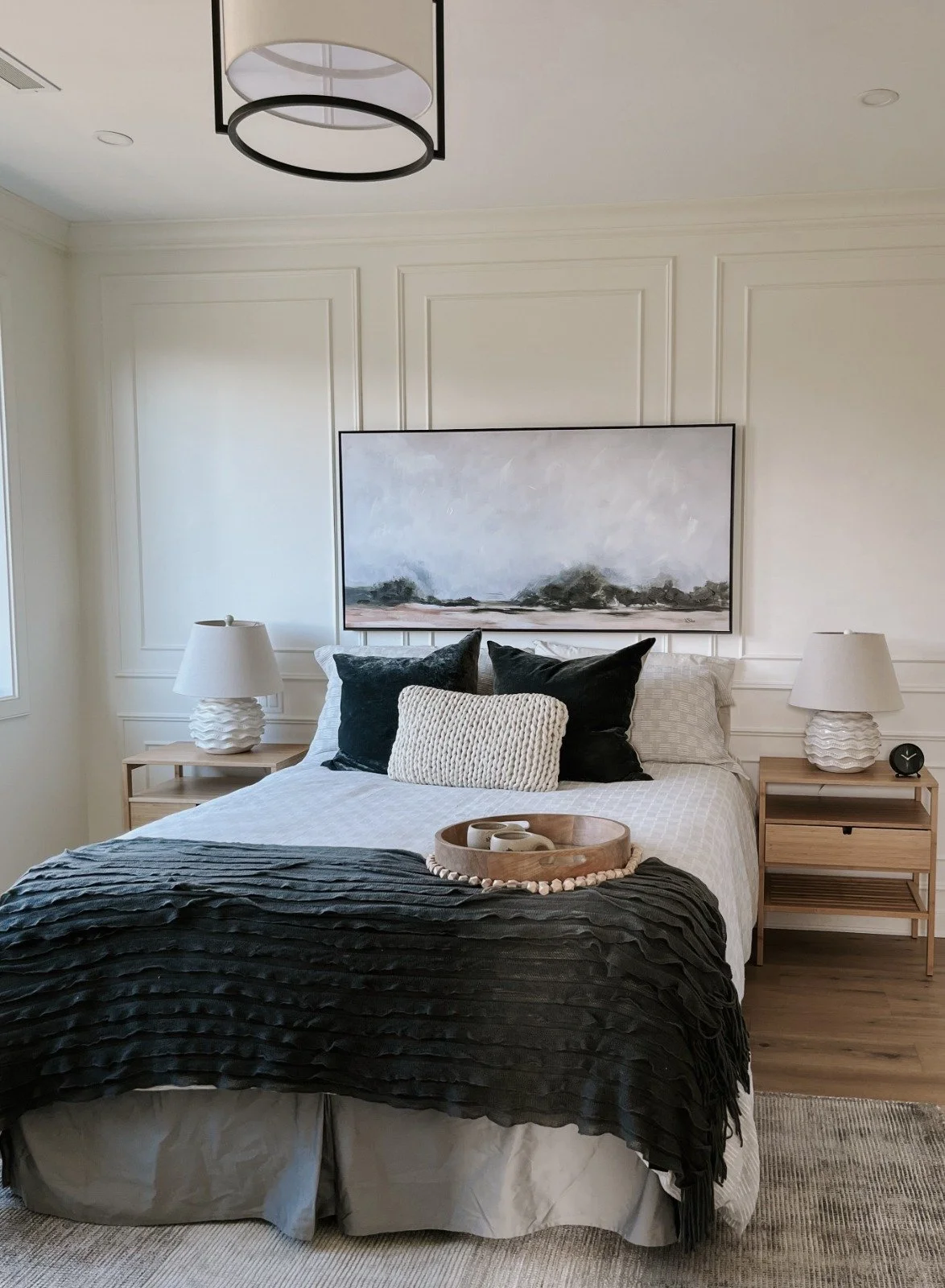 A neatly made bed with black, white, and neutral pillows, a black textured blanket, and a wooden tray with mugs. Two bedside tables with white lamps and a small black clock on the right table. Above the bed is a large abstract landscape painting. The