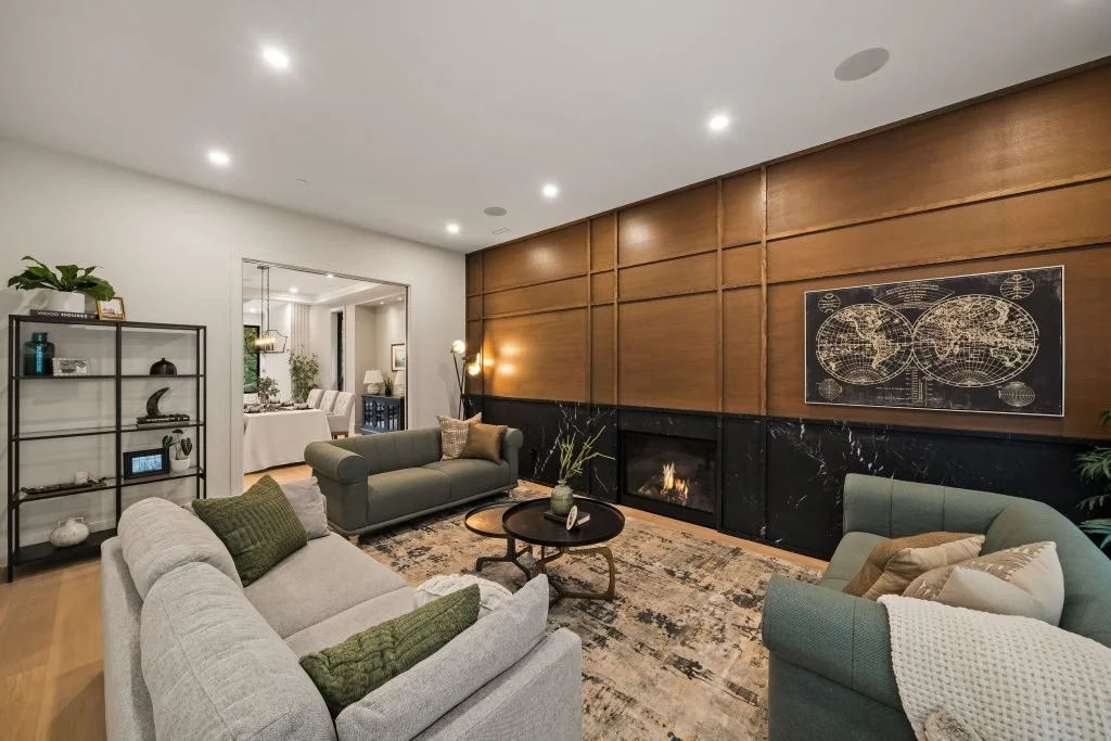 Modern living room with gray and green sofas, a black marble fireplace, wooden wall paneling, a world map art piece, and a patterned rug, with an open dining area in the background.