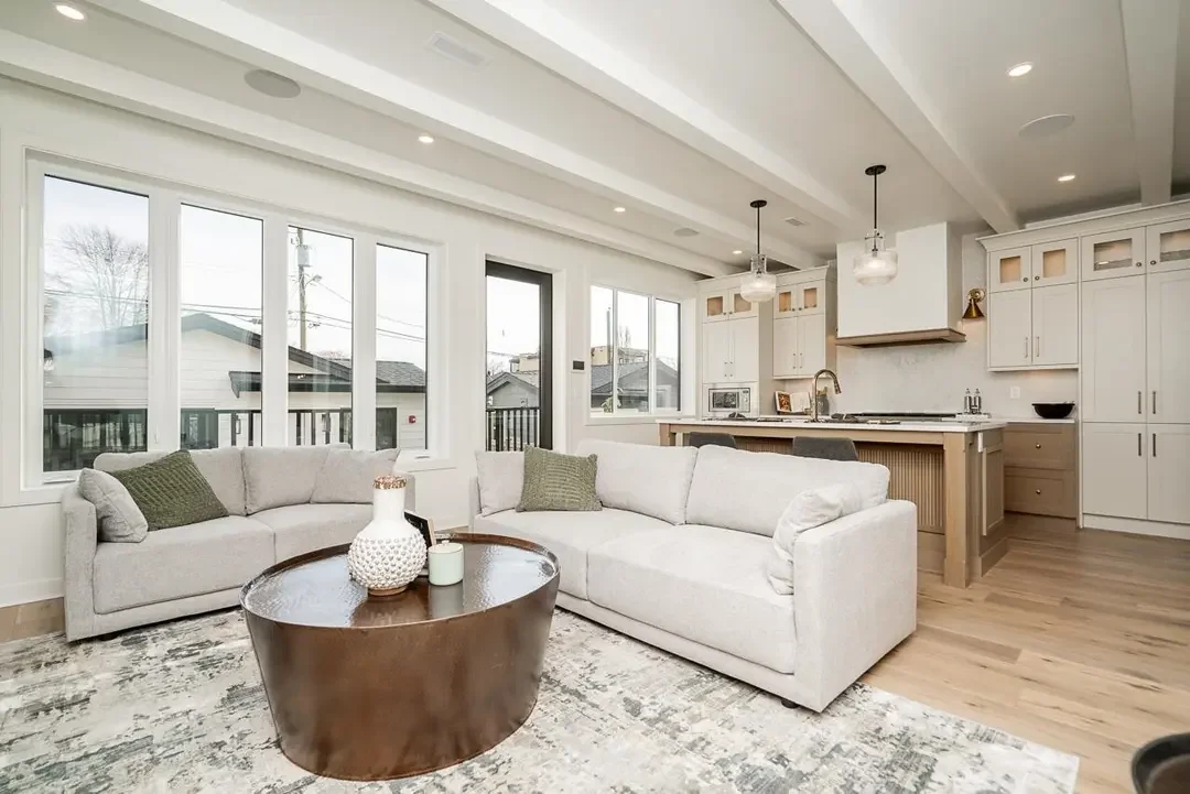 Bright open-concept living room and kitchen with white walls, large windows, beige sofas, a round wooden coffee table, pendant lights, and white cabinetry in the kitchen.