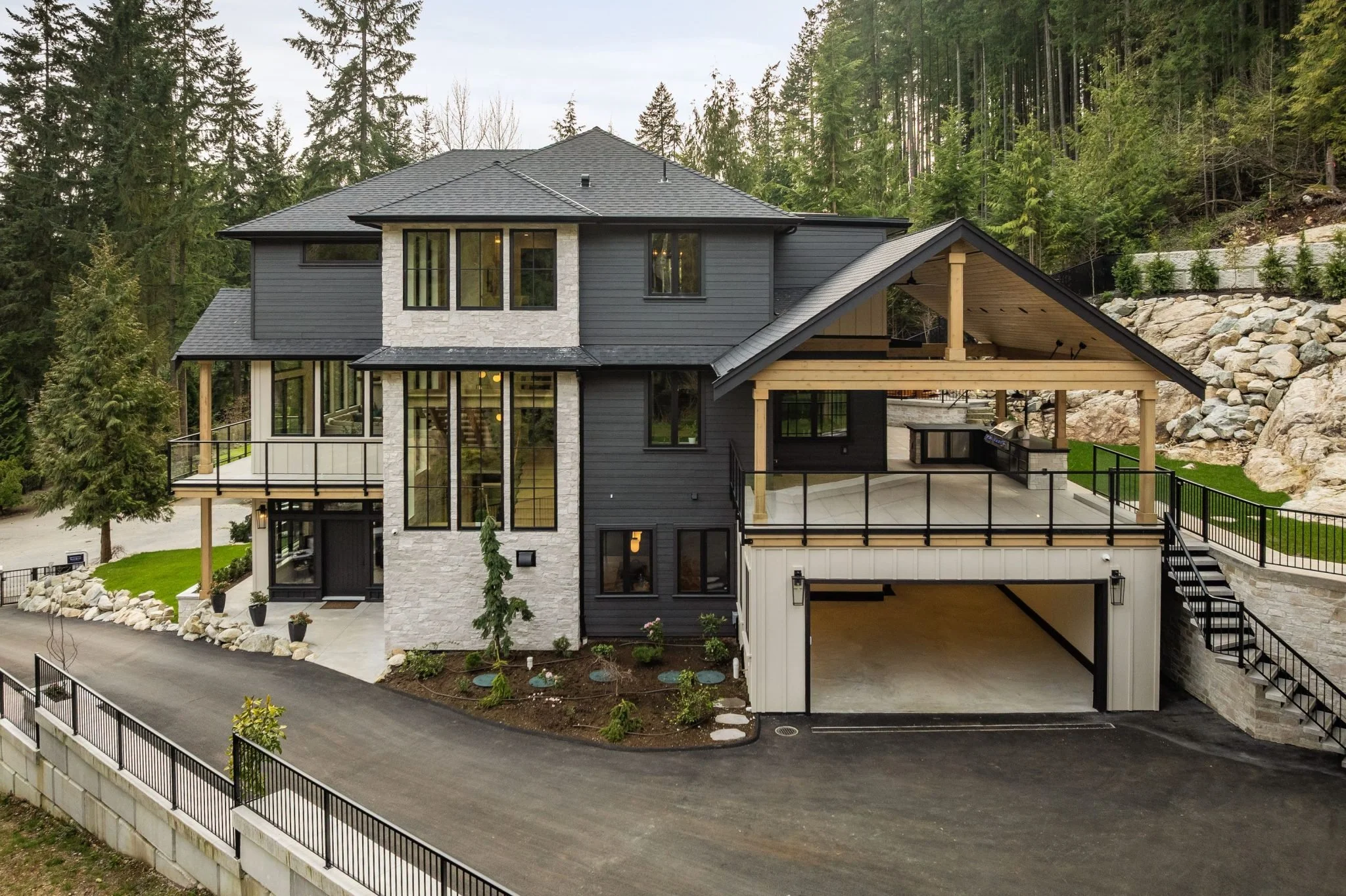 Modern multi-story house with black siding, stone accents, large windows, a spacious covered deck, and an attached garage, surrounded by trees and rocks.