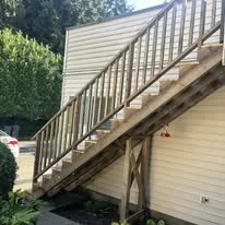 Outdoor wooden staircase attached to a house with beige siding, leading to an upper level, with trees and a white car partially visible in the background.