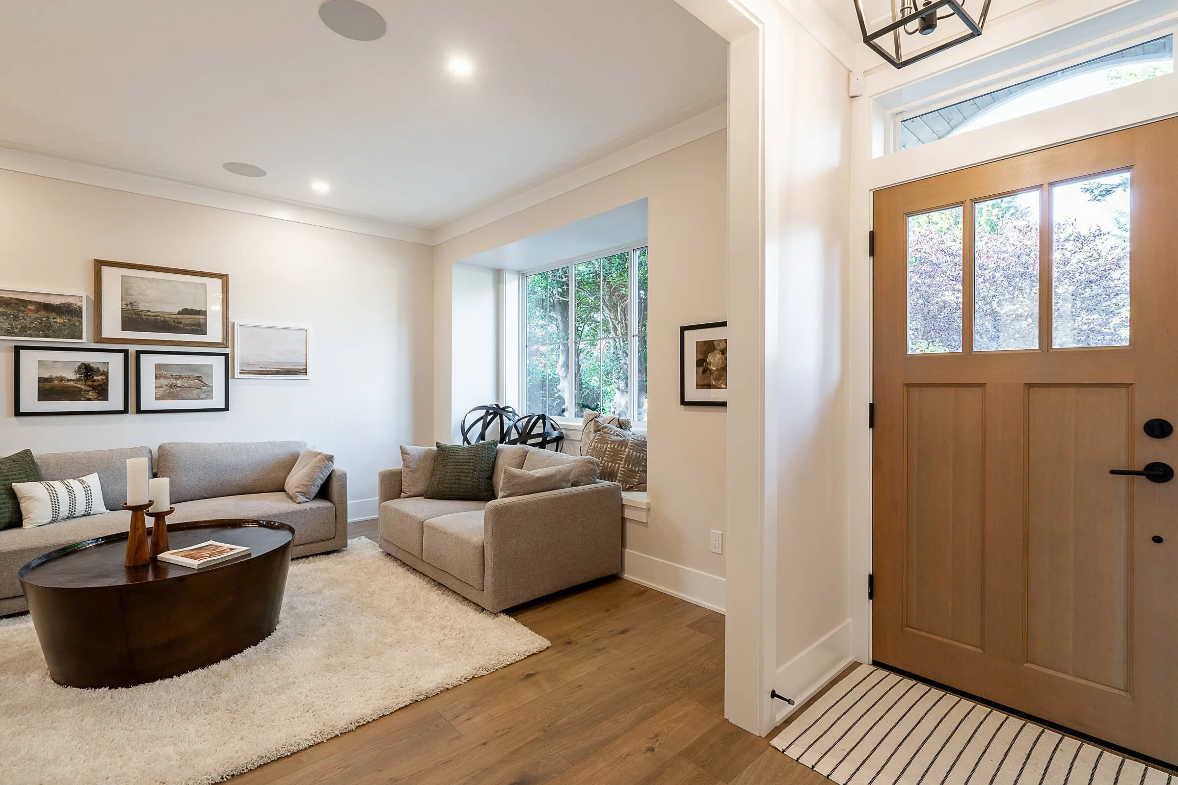 The image shows a cozy and modern living room with a wooden front door on the right and a bright window on the left. The room features beige sofas with green and neutral cushions, a round wooden coffee table, framed landscape artwork on the white wal