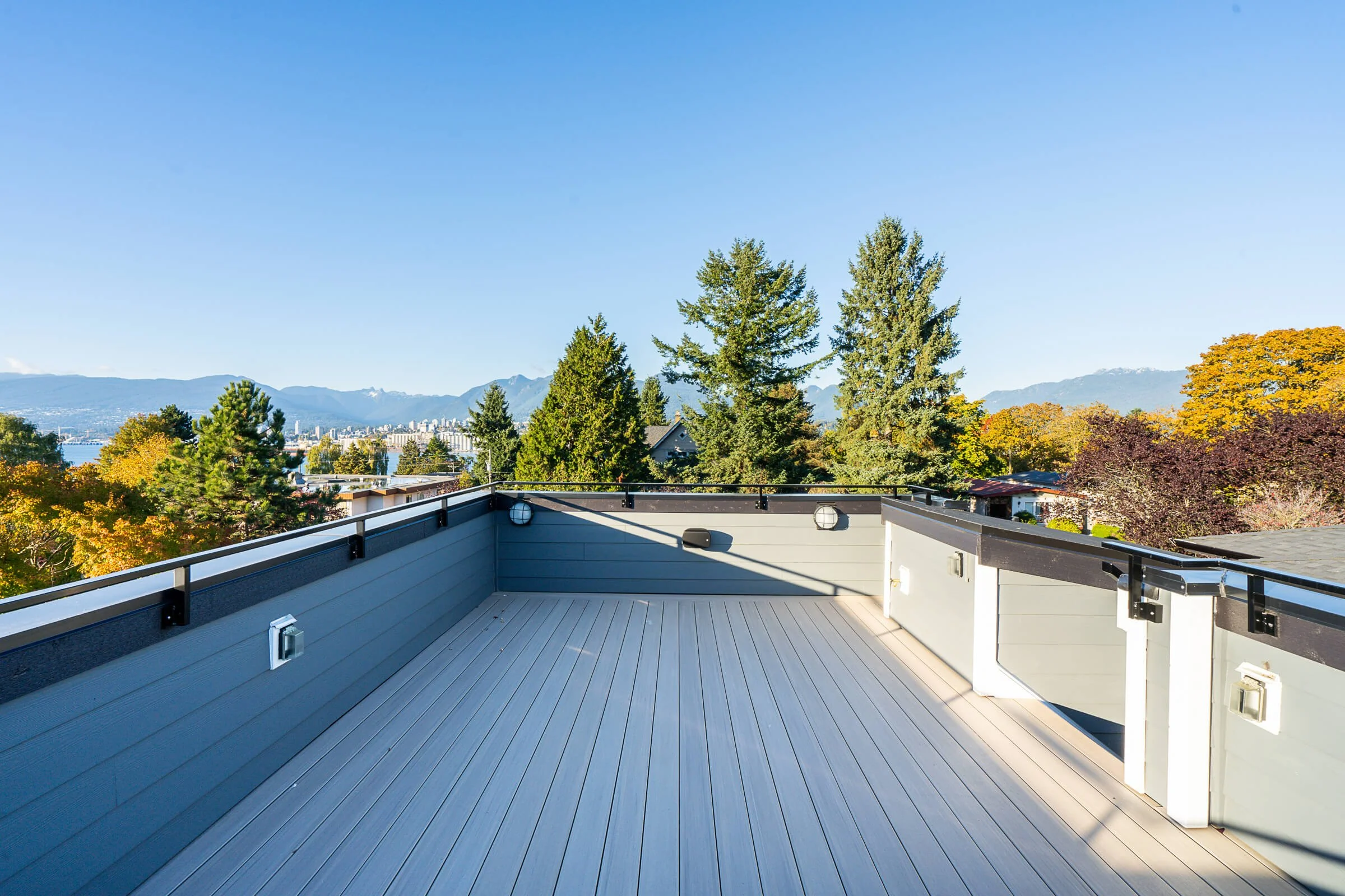 Empty rooftop deck with gray flooring, black railing, and a scenic view of trees, distant city skyline, and mountains under a clear blue sky.