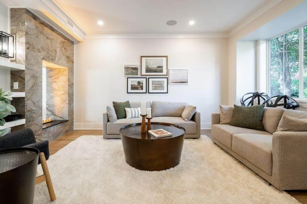 Living room with two beige couches, a round black coffee table, a white shag rug, wall art, a fireplace with a stone surround, and large windows showing greenery outside.
