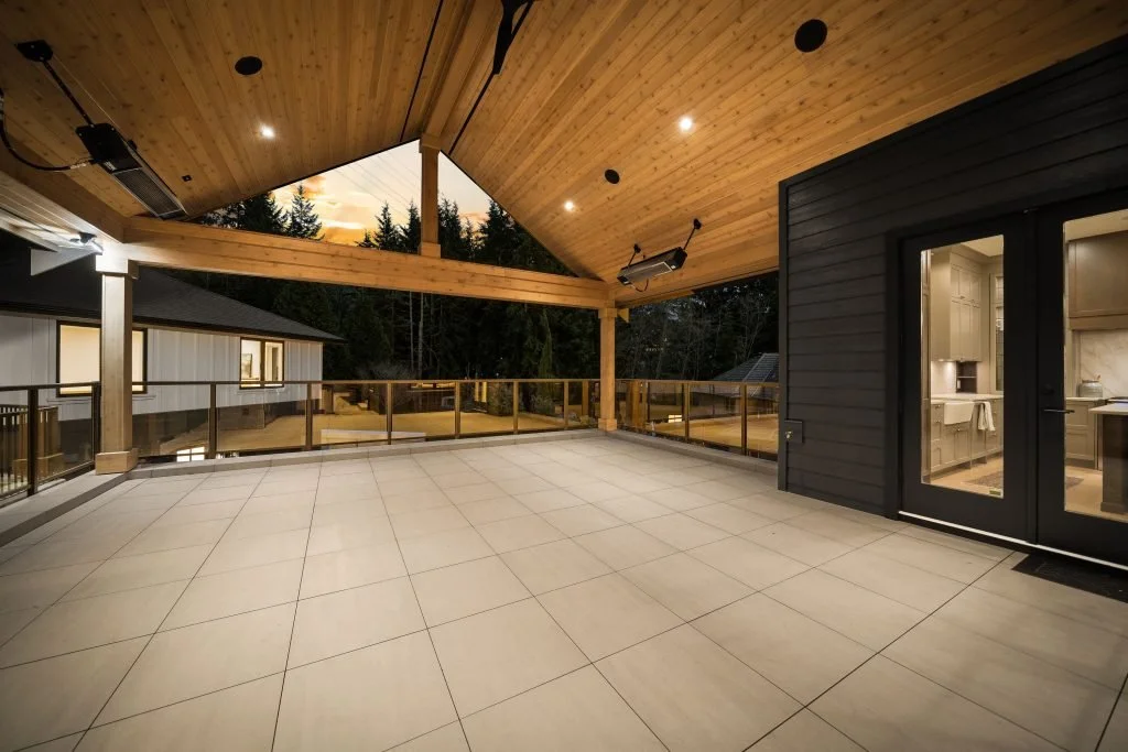 A spacious outdoor balcony with large beige tiles, wooden ceiling, and glass railing, attached to a house with a view of trees and neighboring homes at sunset.