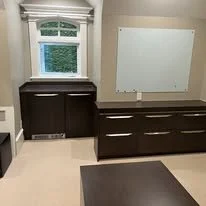 A room with a window, white walls, and dark wood furniture, including a large dresser and a matching cabinet, with a small portion of a dark table visible.
