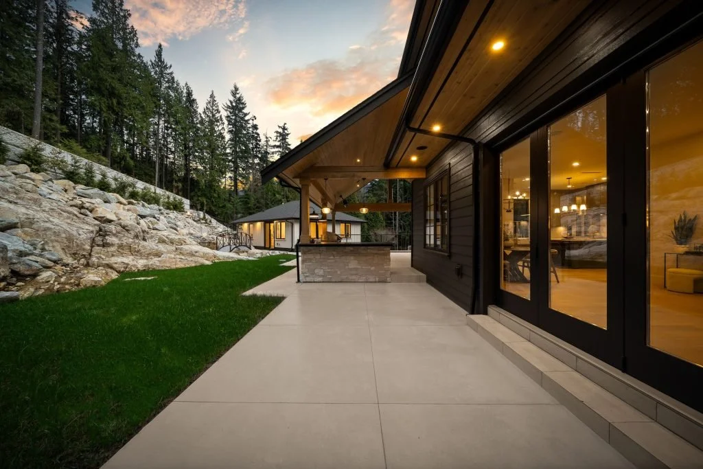A modern house with large glass doors and a covered patio, surrounded by a grassy yard, rocky hillside, and tall trees, during sunset.