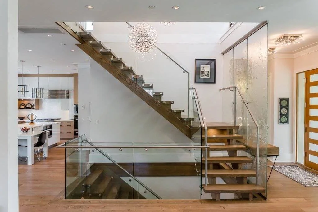 Modern home interior with wooden staircase, glass railing, and adjacent kitchen area.