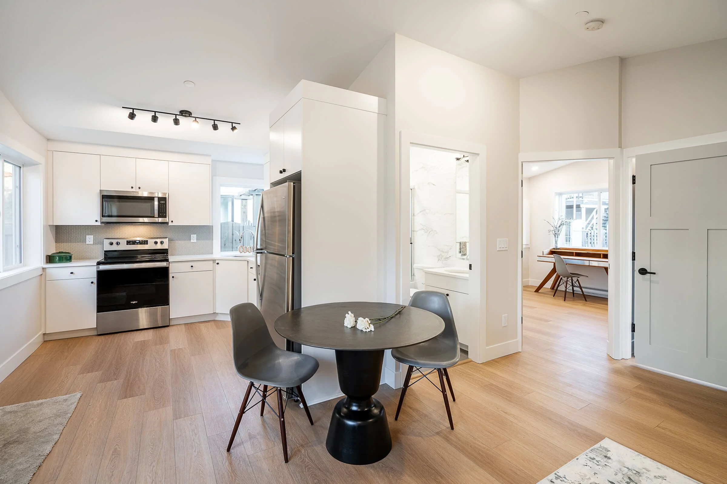 Modern open-plan kitchen and dining area with white cabinets, stainless steel appliances, a round black dining table with three gray chairs, and light wood flooring.
