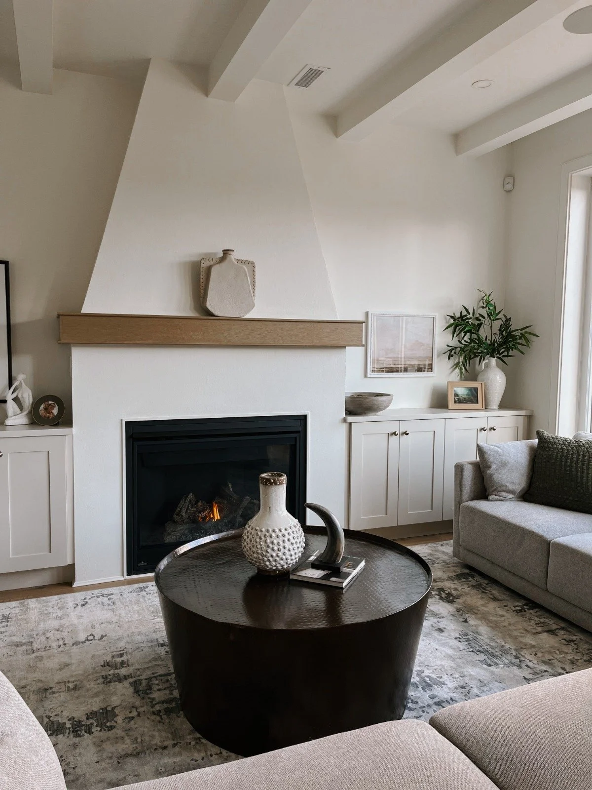 Living room with a white fireplace, gray sofa, black coffee table with decorative vases, a built-in cabinet with framed photos and greenery, and large windows letting in natural light.