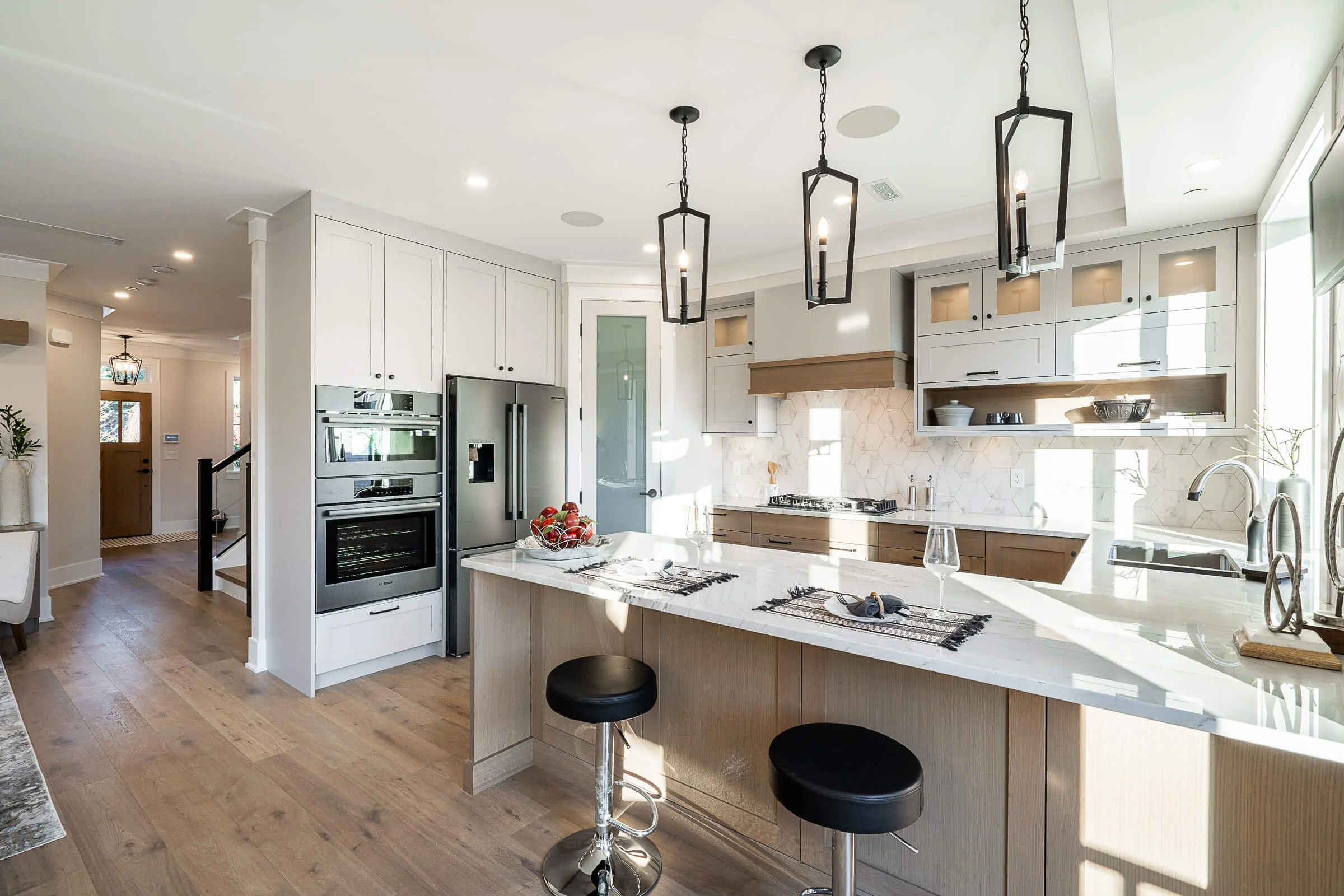 Modern kitchen with white cabinets, stainless steel appliances, a marble island with two black stools, and black pendant lights.