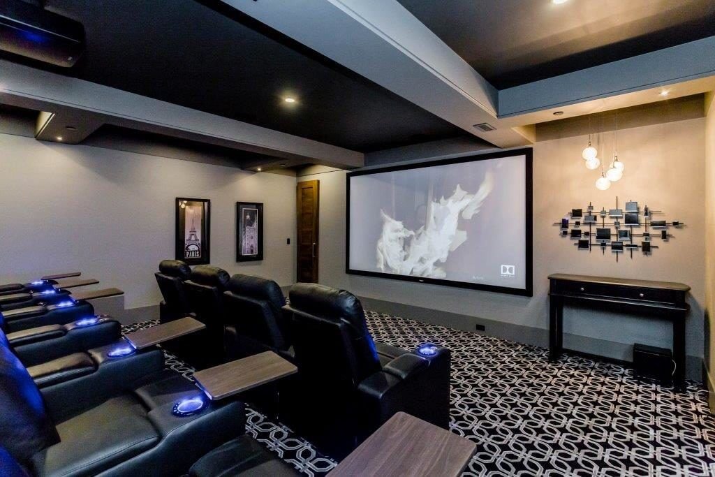 Home theater room with black leather recliners, small side tables, a large projector screen showing a movie, patterned carpet, wall art, and modern lighting.