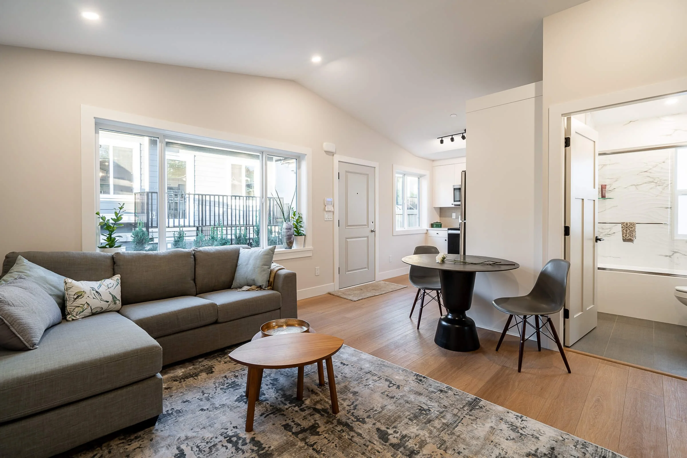 Living room with a gray sofa, a small wooden coffee table, large windows with plants, a round black dining table with chairs, and a view into the kitchen and bathroom with a shower.