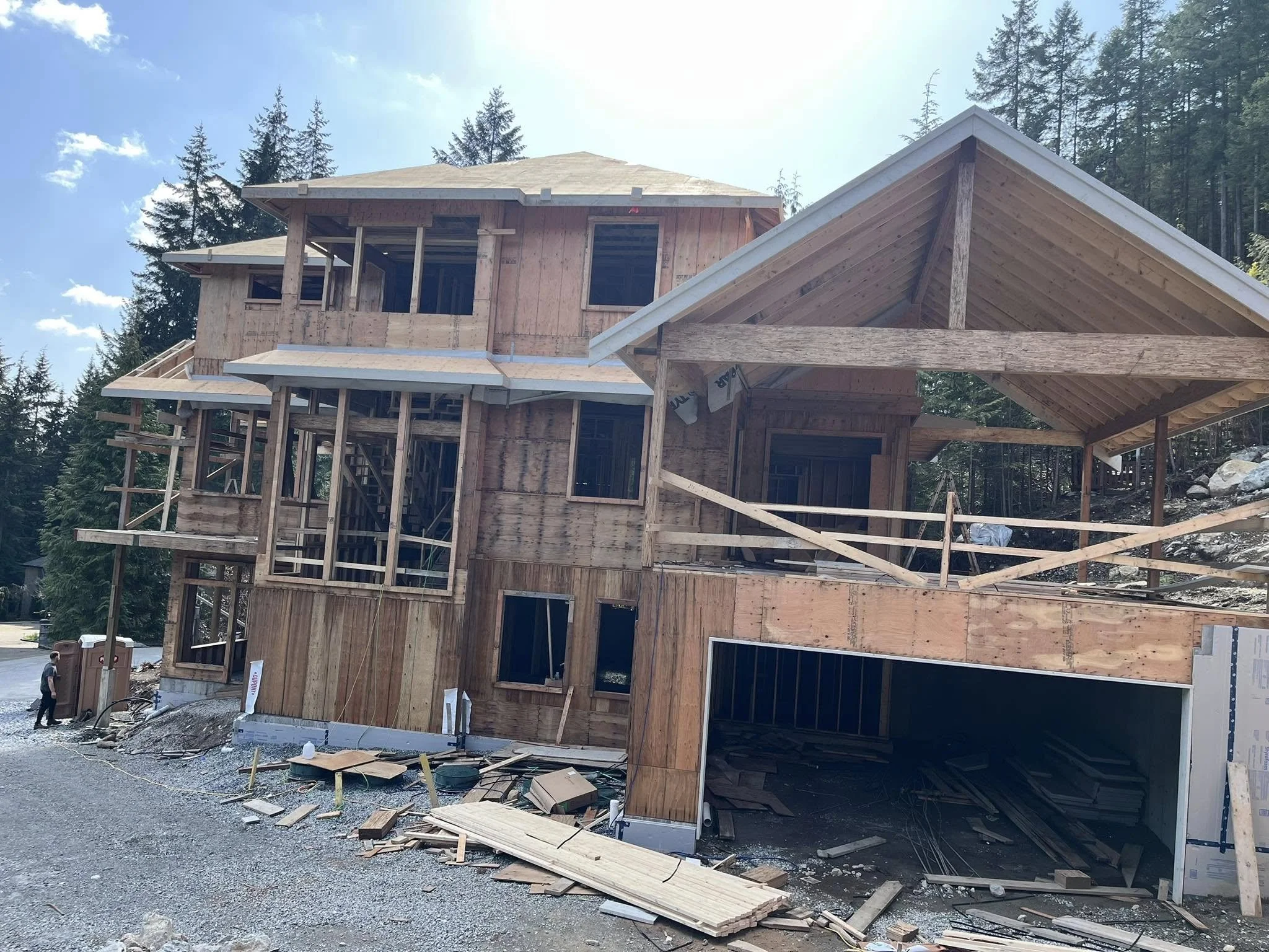 A multi-story house under construction surrounded by trees, with exposed wooden framing, uninstalled windows, and building materials scattered around.
