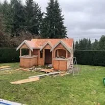 A small wooden house under construction in a grassy yard with trees in the background, construction materials scattered around.