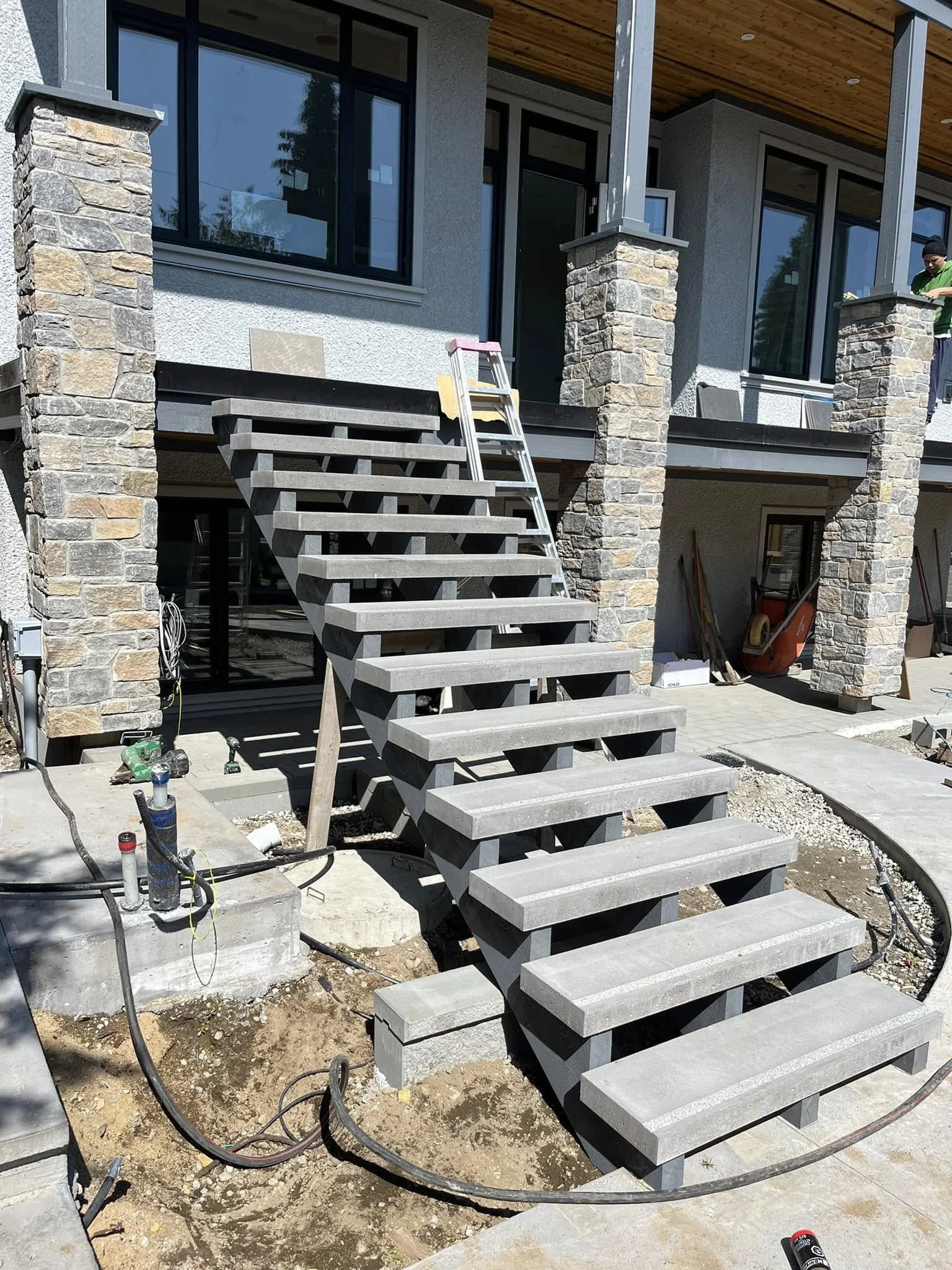Construction site showing an unfinished outdoor staircase leading to a house with stone columns and large windows; construction tools and materials are visible around.