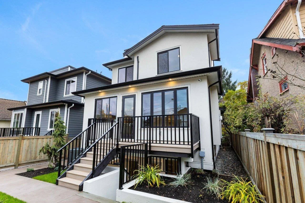 Modern white multi-story house with black railings and large glass windows, front steps leading to a porch, and a small landscaped yard with plants and grass.