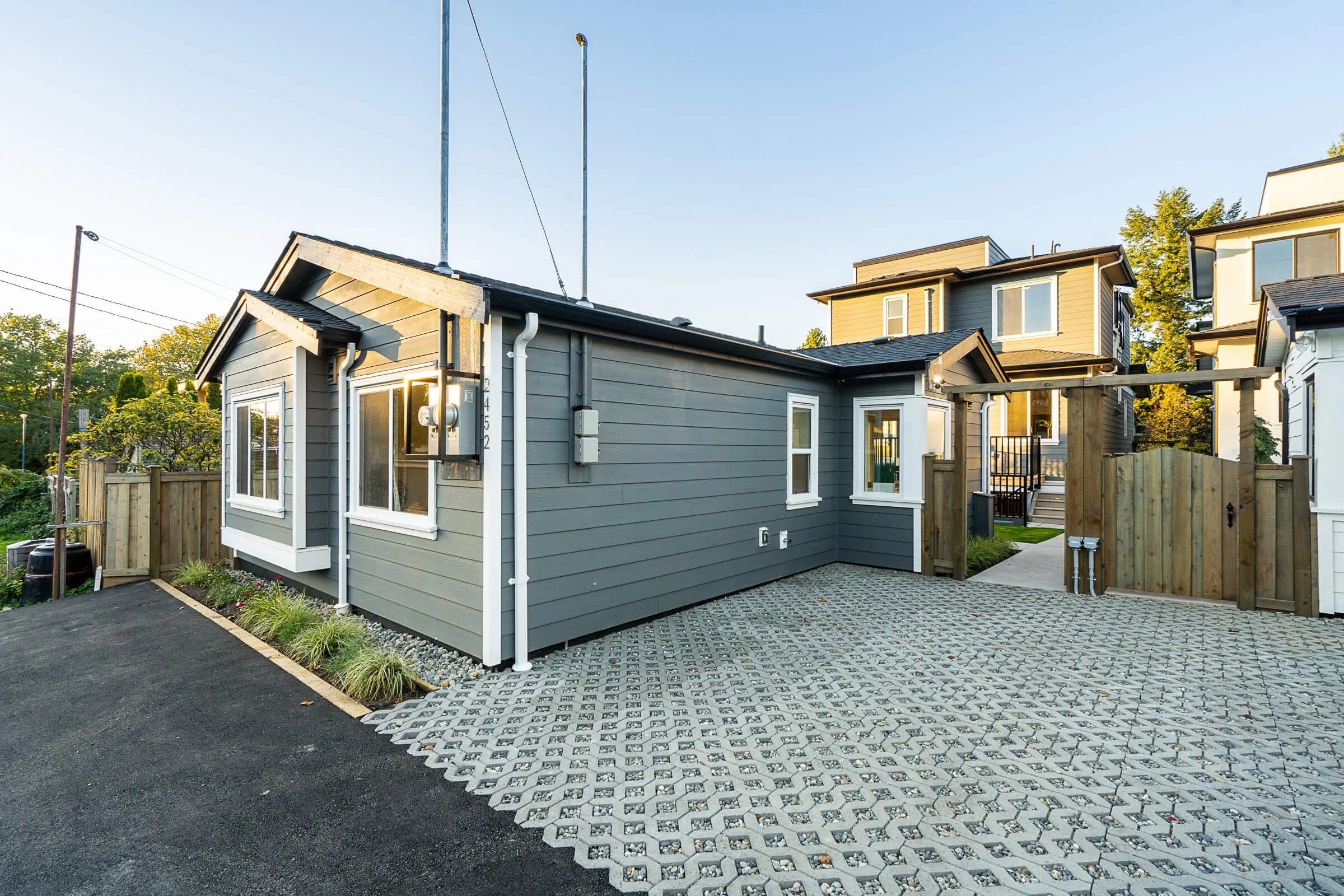 Front view of a modern house with gray siding, white trim, and a small porch, surrounded by a wooden fence and paving stones in the driveway during daylight.