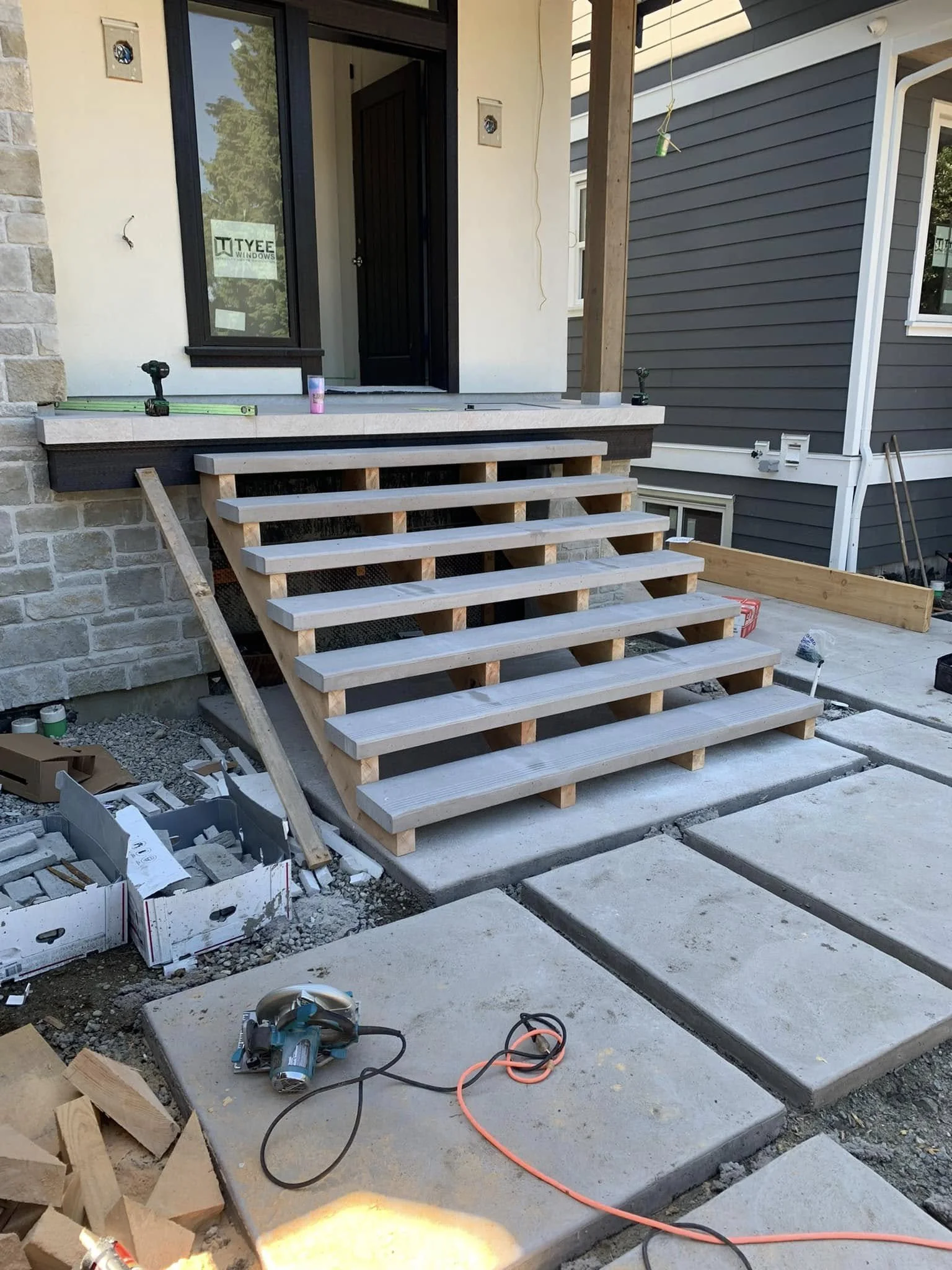 Construction site with wooden stairs leading to a front porch, tools and building materials scattered around, including a power sander, extension cord, and concrete slabs.