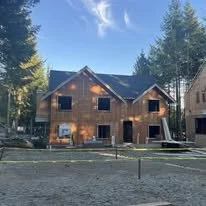 A two-story house under construction with wooden exterior, surrounded by trees and a clear sky.