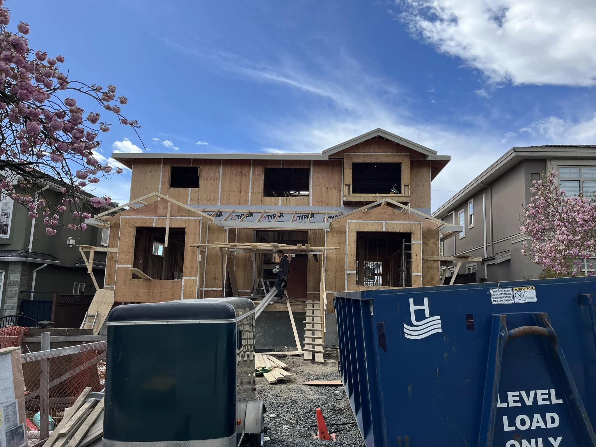 A two-story house under construction with wooden framing, scaffolding, and construction workers working inside, surrounded by construction equipment and materials, alongside neighboring finished houses and cherry blossom trees under a partly cloudy s