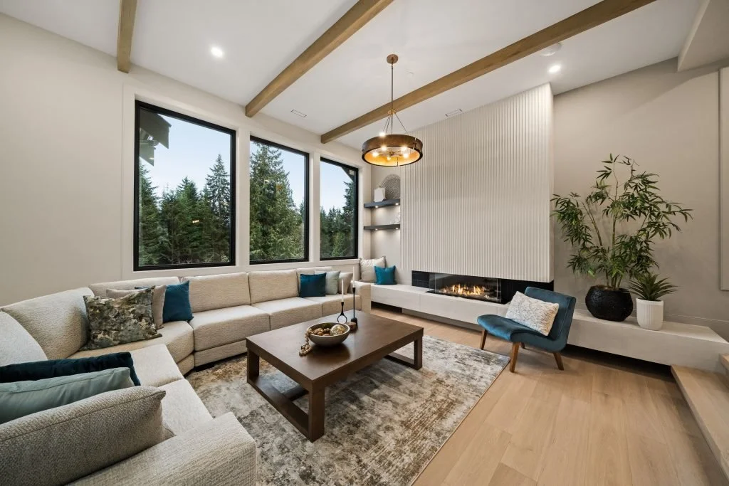 Modern living room with large windows overlooking trees, beige sofa with colorful pillows, a wooden coffee table, a blue chair, built-in white TV console with a fireplace, large potted plant, wood beams on ceiling, and hardwood flooring.