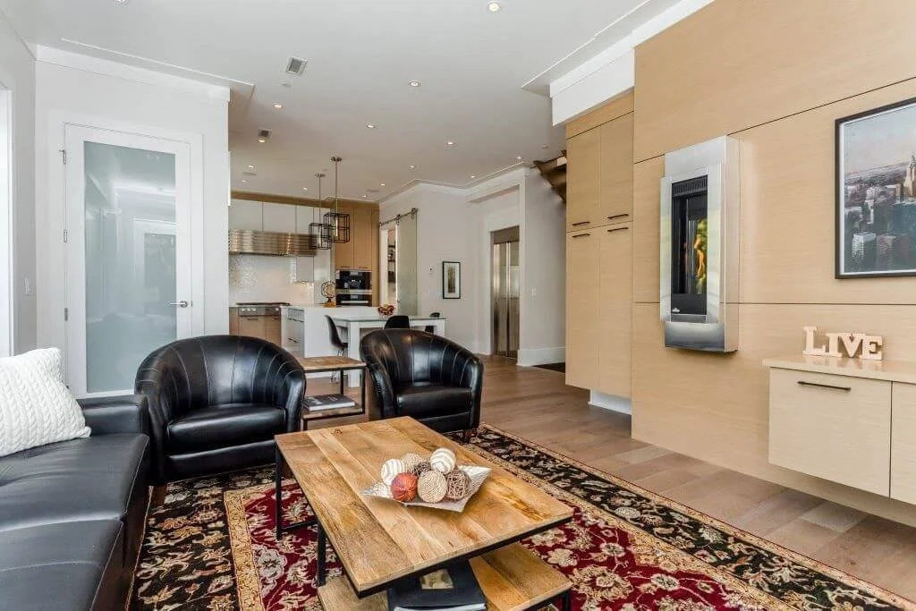 Modern living room with black leather sofa and chairs, wooden coffee table, area rug, and open kitchen in the background.