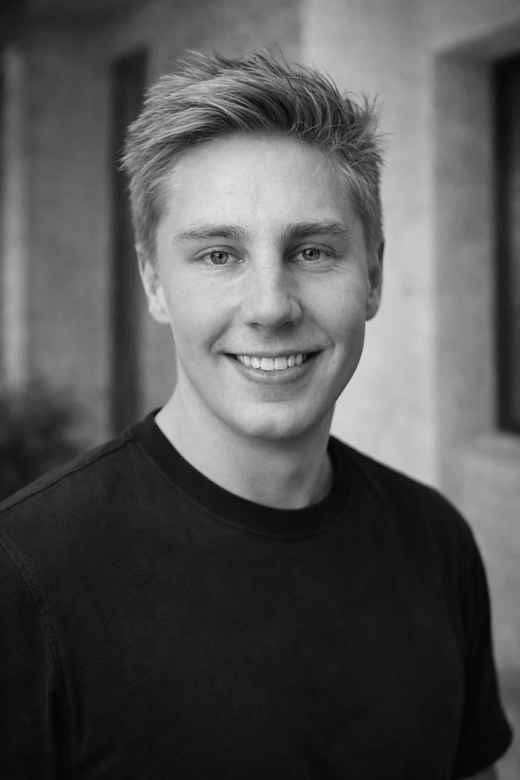 Black and white portrait of a young man with short, styled hair, smiling, wearing a black T-shirt, outdoors near a brick building.