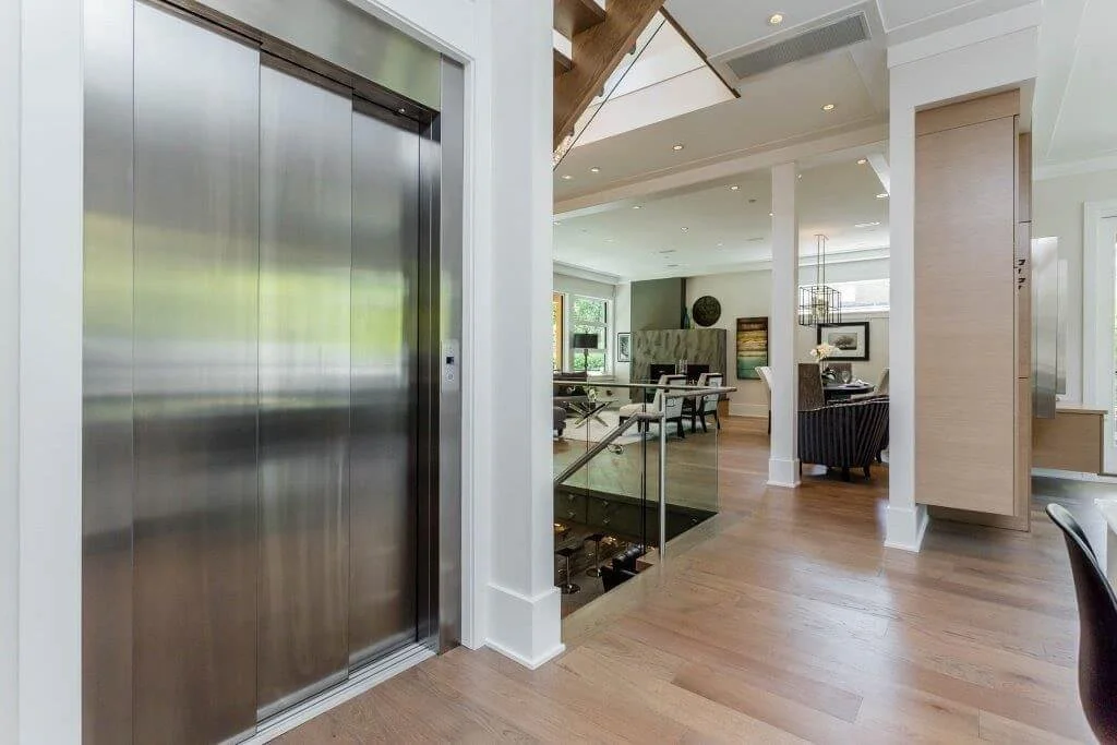 Modern residential interior with a stainless steel elevator door, open plan living and dining area, wooden flooring, and large windows
