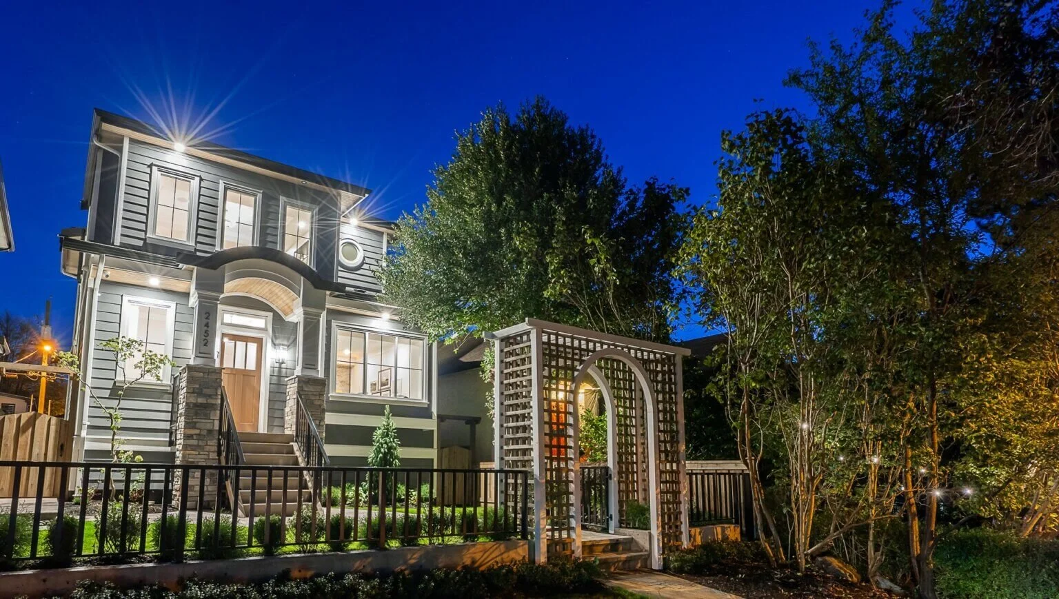A well-lit, two-story house at night with exterior lights, a front porch, a staircase, and a garden with trees and a decorative archway in front.