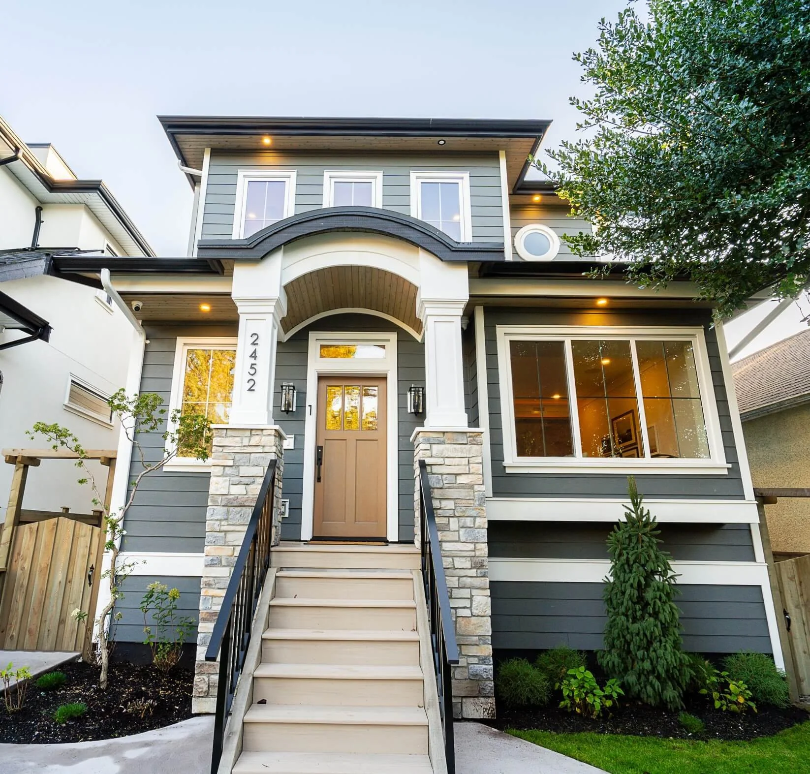 A modern two-story house with dark gray siding, white trim, stone accents on the front pillars, and a beige front door. Wide front steps lead up to the entrance, with small trees and bushes in the landscaped yard.