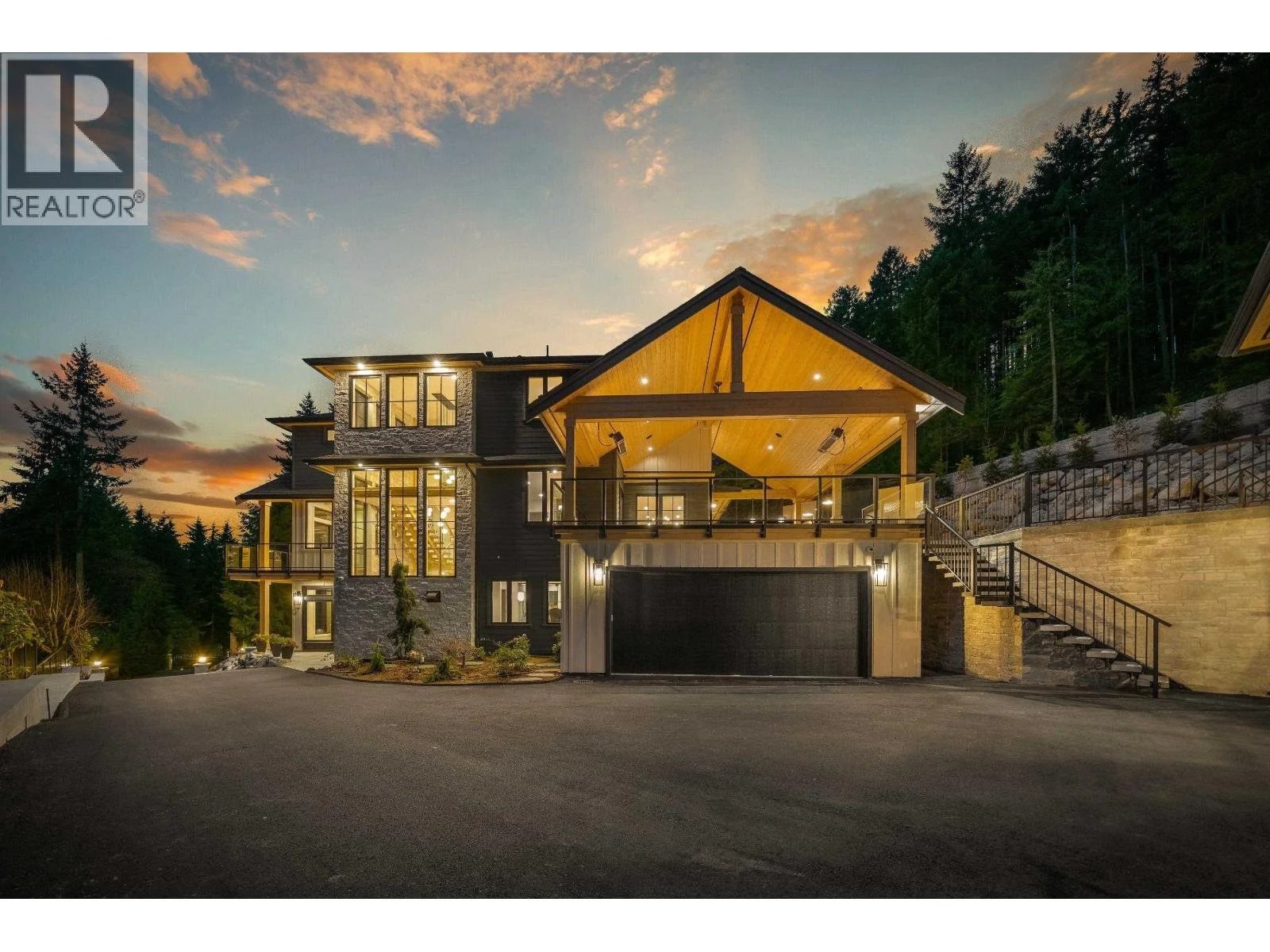 Modern multi-story house with large glass windows, stone and dark wood exterior, lit interior, situated on a hillside with trees, at dusk with a colorful sky.