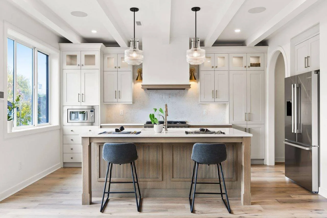 Modern kitchen with white cabinets, a central island with two dark gray chairs, a stainless steel refrigerator, a microwave, and two pendant lights hanging above the island, with natural light coming through a window on the left.