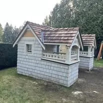 Small wooden playhouse with a shingled roof, front porch, and window, situated in a backyard with trees and a fence in the background.