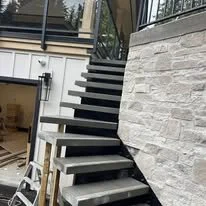 Exterior staircase with black steps and white risers, adjacent to a stone wall and glass railing, under construction or renovation.