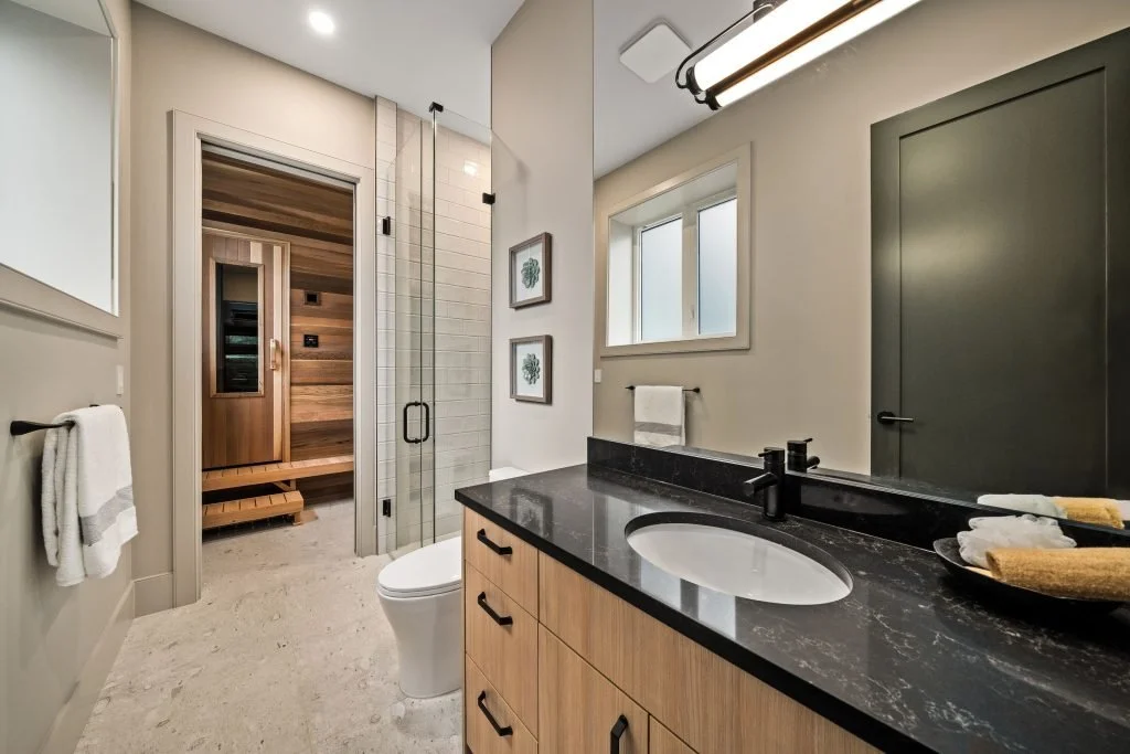 Modern bathroom with a black countertop vanity, a white oval sink, black fixtures, and a large mirror. There is a window with frosted glass, a towel rack, and a glass shower enclosure. A wooden sauna is visible through the open door.