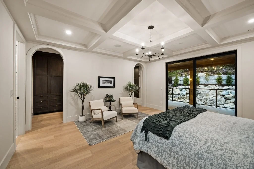 A bedroom with a coffered ceiling, a chandelier, a large sliding glass door to a patio, a bed with a dark throw blanket, and a seating area with two chairs, potted plants, and a framed picture on the wall.
