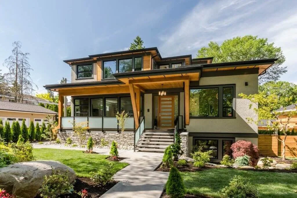 Modern two-story house with large windows, a front porch, and landscaped garden with trees and shrubs.