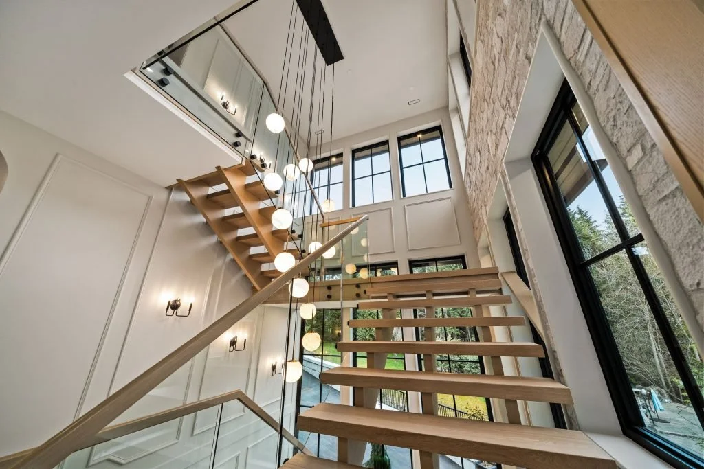 Modern wooden staircase inside a house with large windows, hanging globe lights, and stone accent wall.